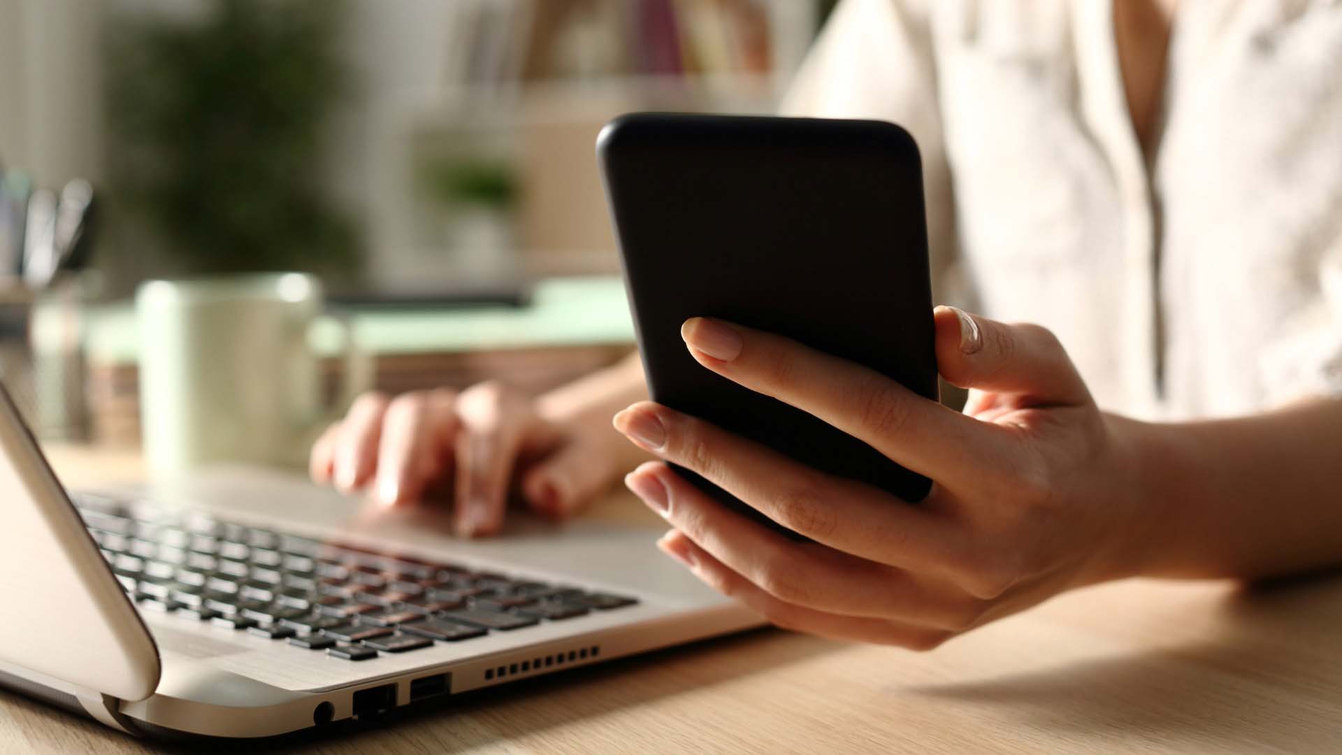 Woman's hand holding smartphone and on laptop simultaneously.